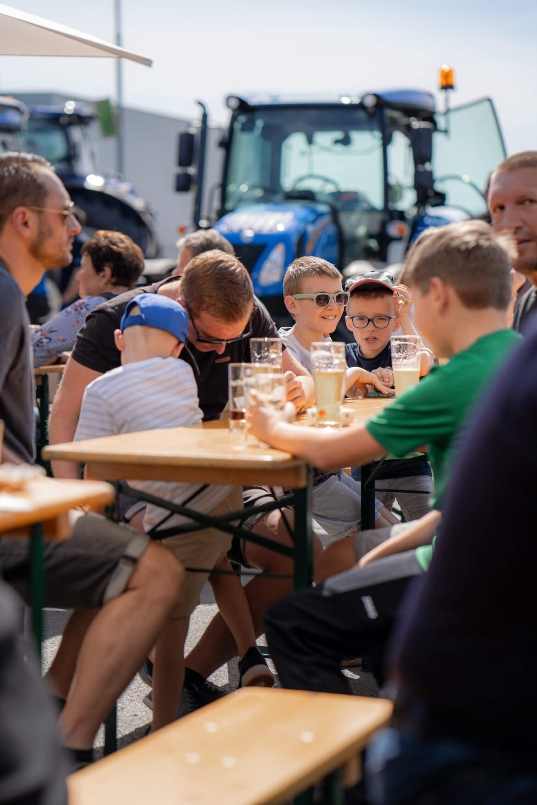 Familienfest im Freien: Erwachsene und Kinder genießen Getränke an einem Biertisch, Traktoren im Hintergrund.