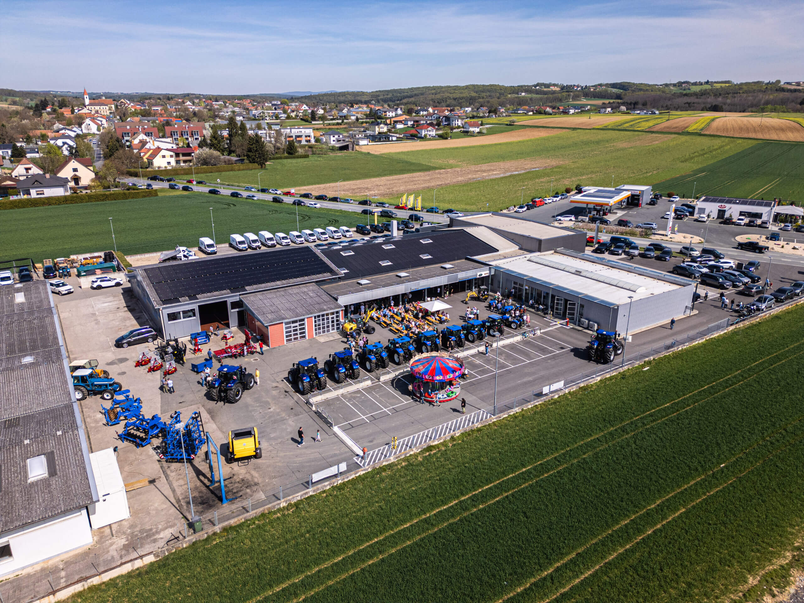 Luftaufnahme einer Landmaschinenmesse mit Traktoren und Besuchern, umgeben von Feldern und einem Dorf im Hintergrund.