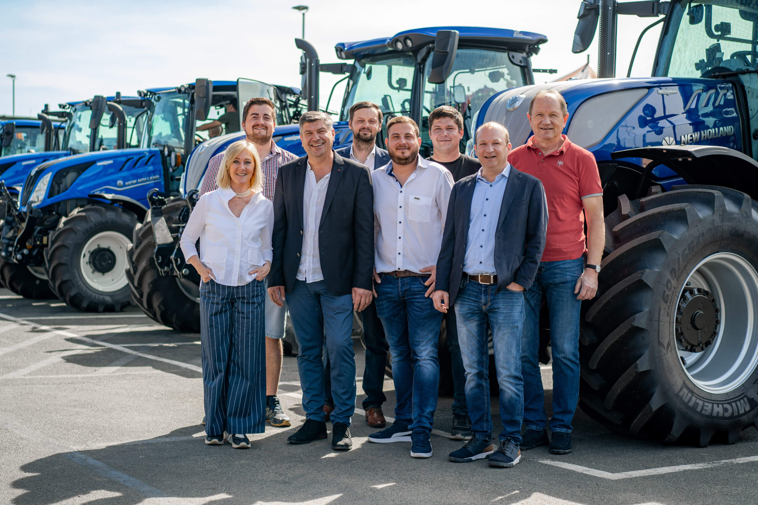 Gruppe von Menschen posiert vor einer Reihe blauer Traktoren bei sonnigem Wetter auf einem Parkplatz.