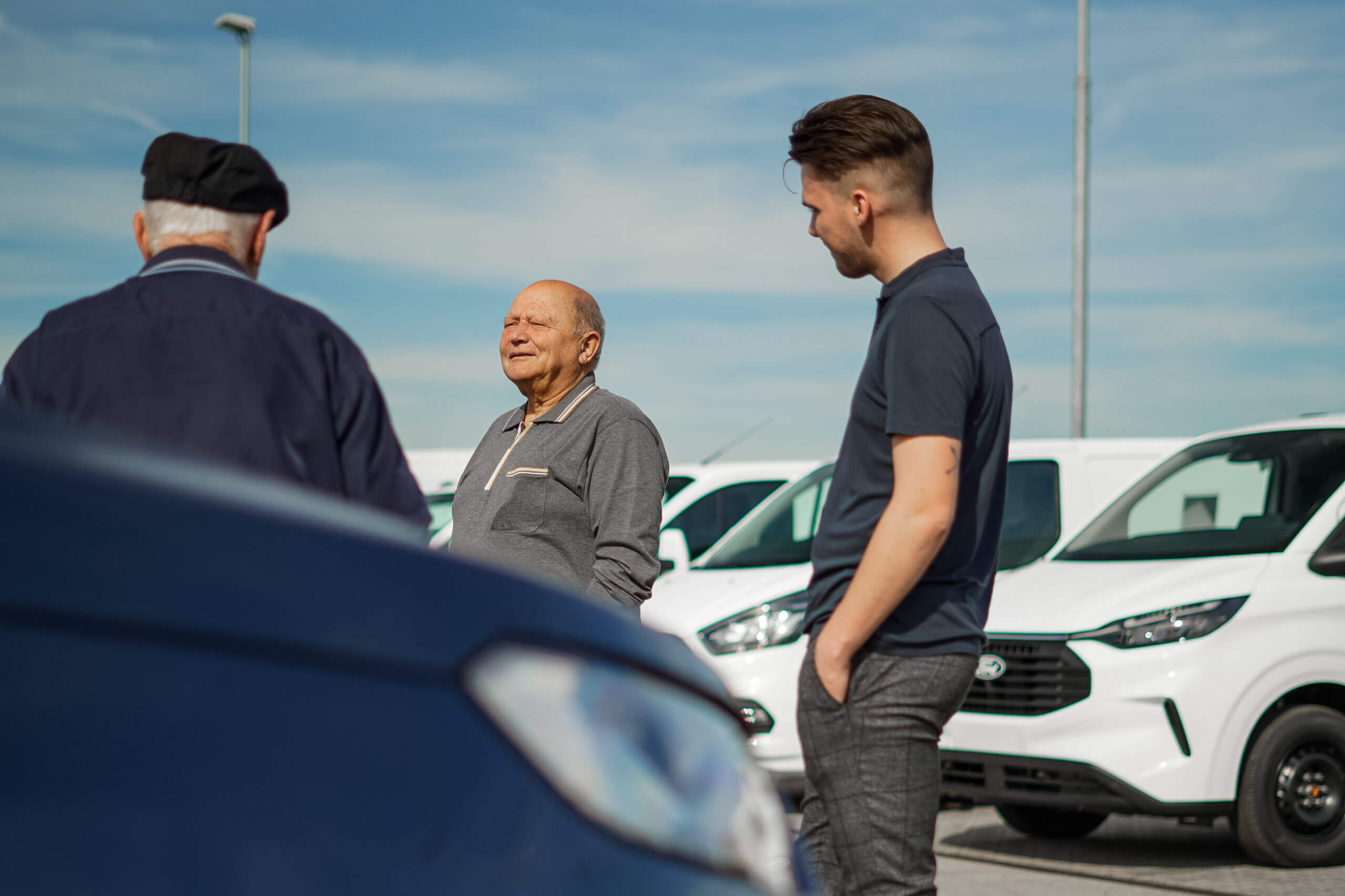 Drei Männer unterhalten sich auf einem Parkplatz, umgeben von weißen Lieferwagen bei sonnigem Wetter.