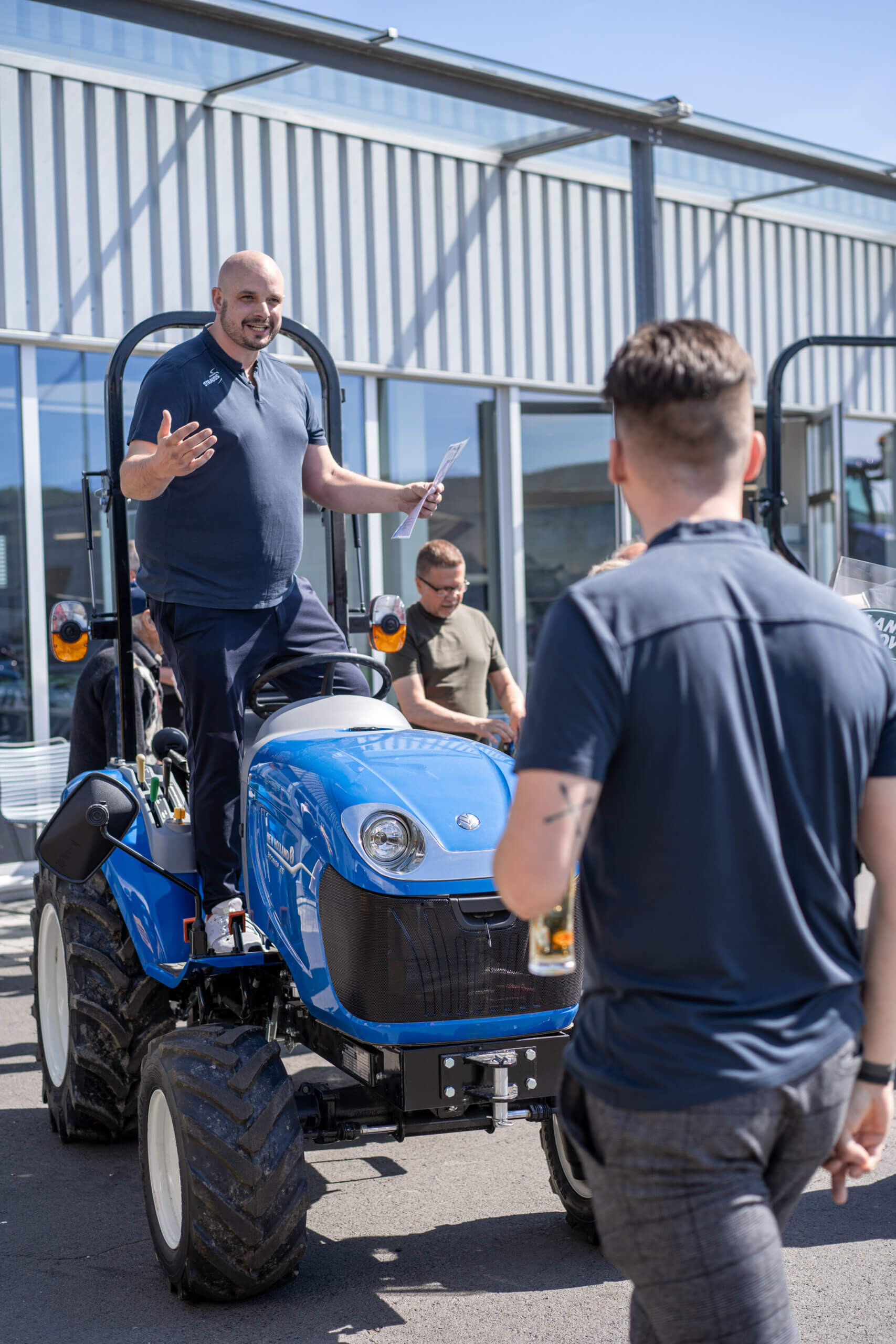 Mann steht auf blauem Traktor und spricht mit Menschen bei sonniger Veranstaltung vor moderner Gebäudefassade.