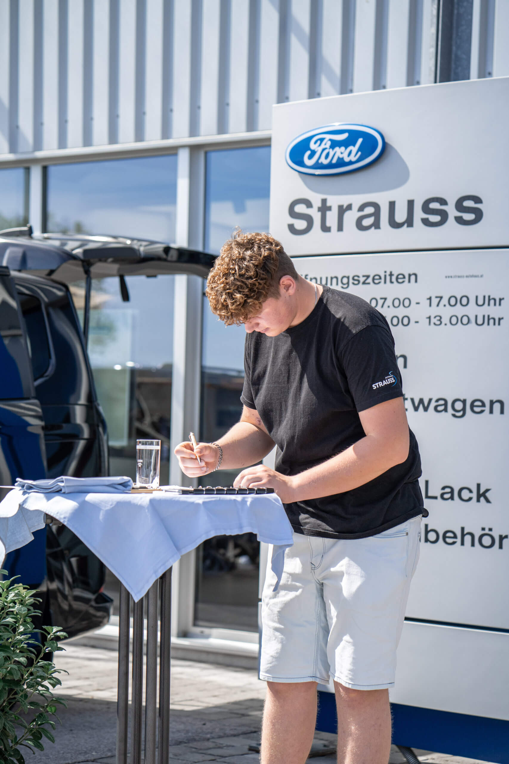 Mann unterschreibt Dokument vor Ford Strauss Autohaus im Freien, nahe einem Tisch mit Glas Wasser und Dokumenten.