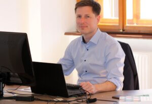 Mann im Büro arbeitet am Computer und lächelt, trägt blaues Hemd, sitzt in sonnigem Raum.uzet gsdfg