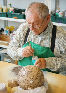 Älterer Mann bemalt Keramikobjekt in Werkstatt, in konzentrierter Handarbeit, mit Pinsel und grünem Schürzenhalter.