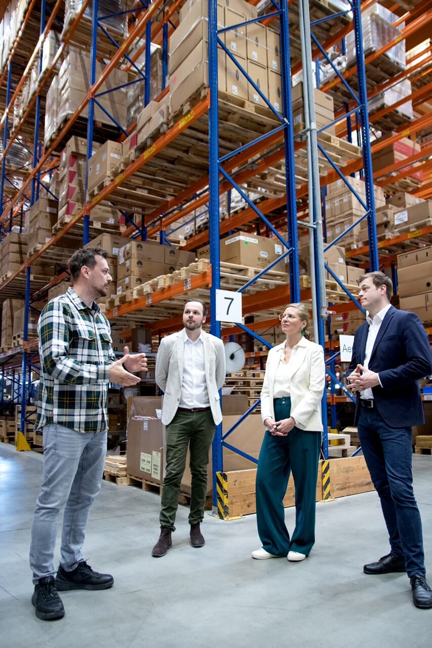 Gruppe diskutiert in einem Lager mit hohen Regalen voller Kartons und Paletten.