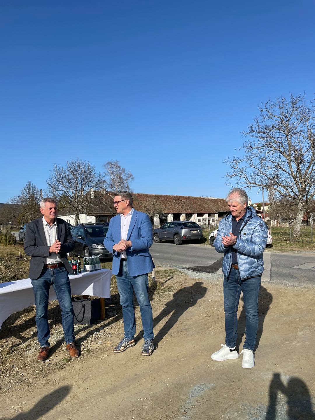 Drei Männer bei einer Outdoor-Veranstaltung auf einem sonnigen Gelände mit Autos im Hintergrund.