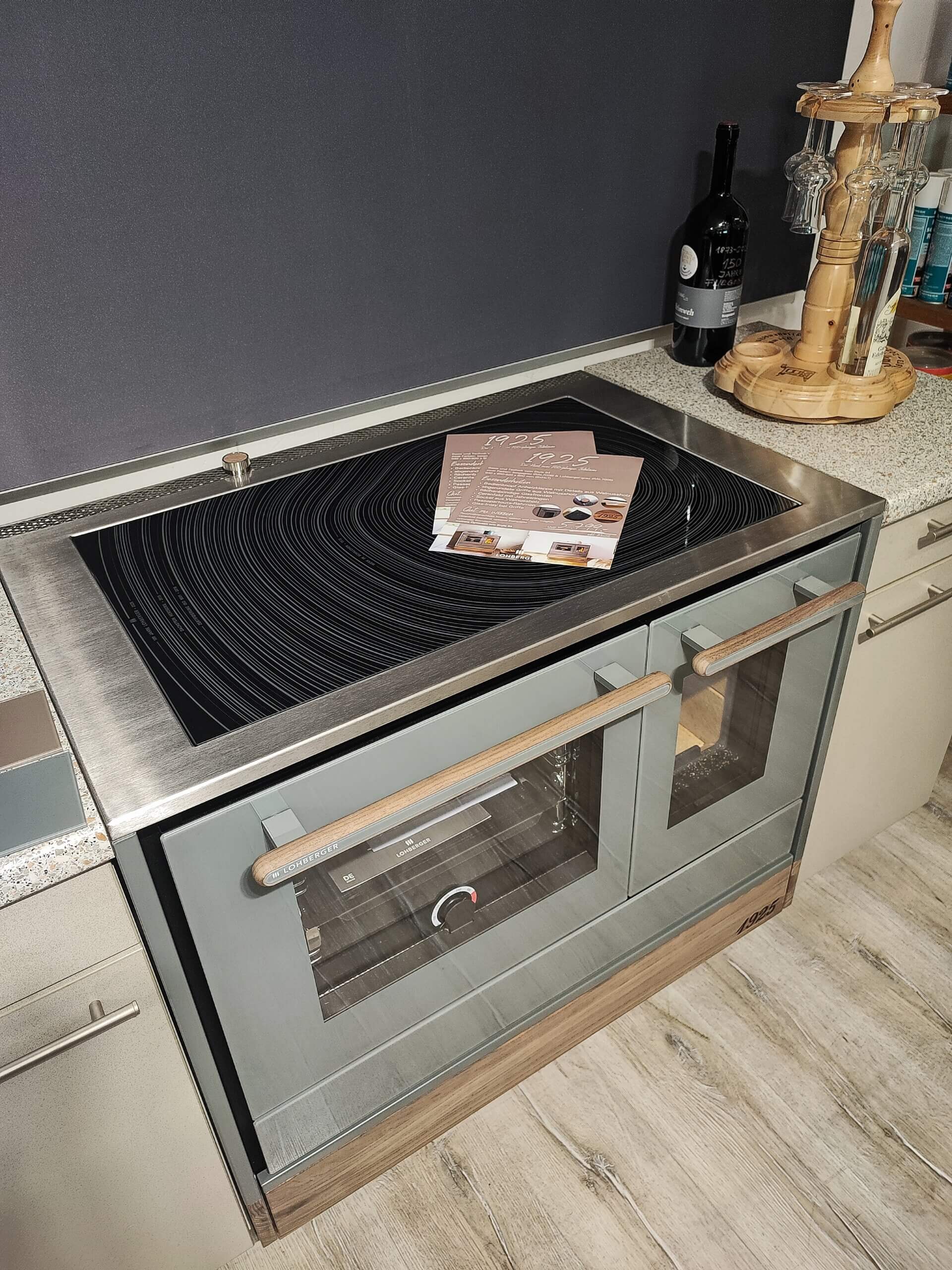 Moderner Küchenherd mit Glaskeramikkochfeld und beige-grauer Front, dekoriert mit Broschüren und einer Weinflasche.
