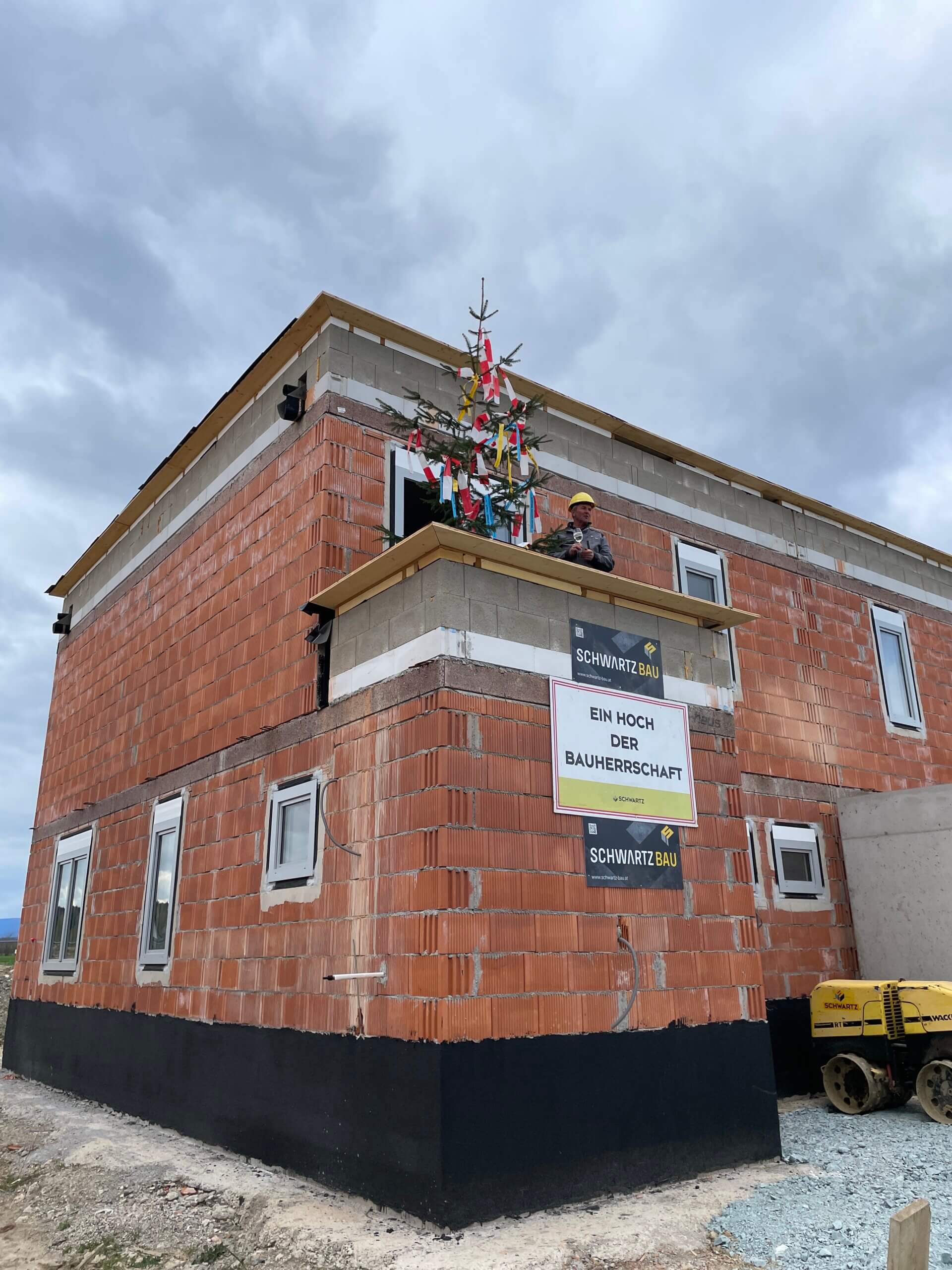 Richtfest bei Rohbau: Geschmückter Baum und Schild „Ein Hoch der Bauherrschaft“ auf zweistöckigem Gebäude.