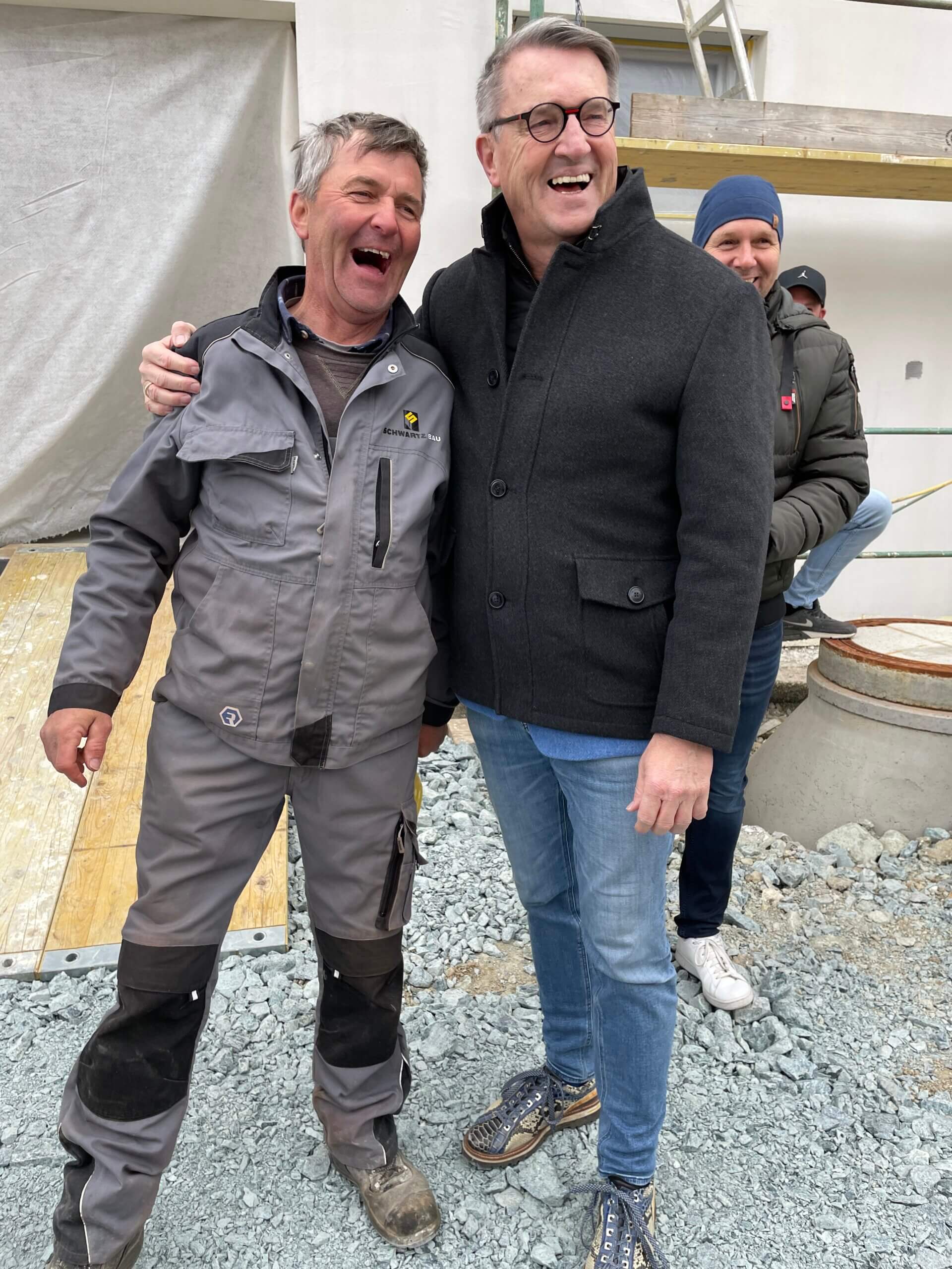 Zwei Männer lachen und umarmen sich auf einer Baustelle, umgeben von Bauschutt und Kollegen im Hintergrund.