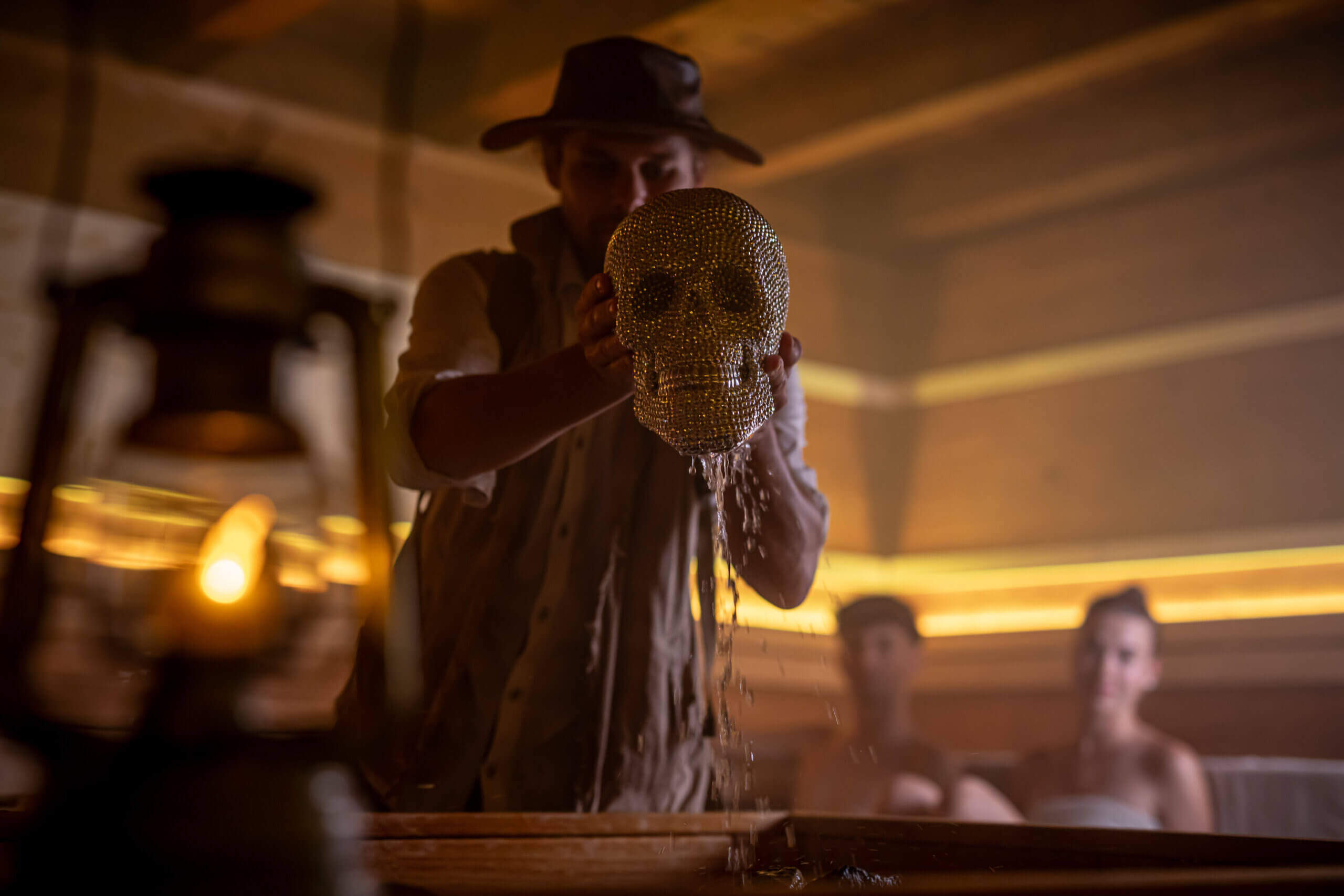 Person in Abenteurerlook hält funkelnden Schädel, während Wasser herunterfließt. Mysteriöser Raum im Hintergrund.