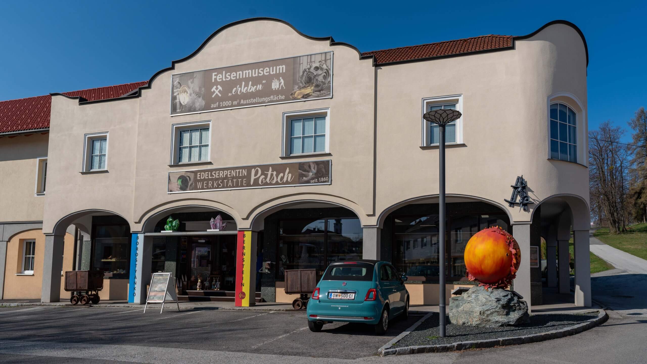 Außenansicht des Felsenmuseums Bernstein mit großem orangenem Kunstobjekt und geparktem Auto an sonnigem Tag.