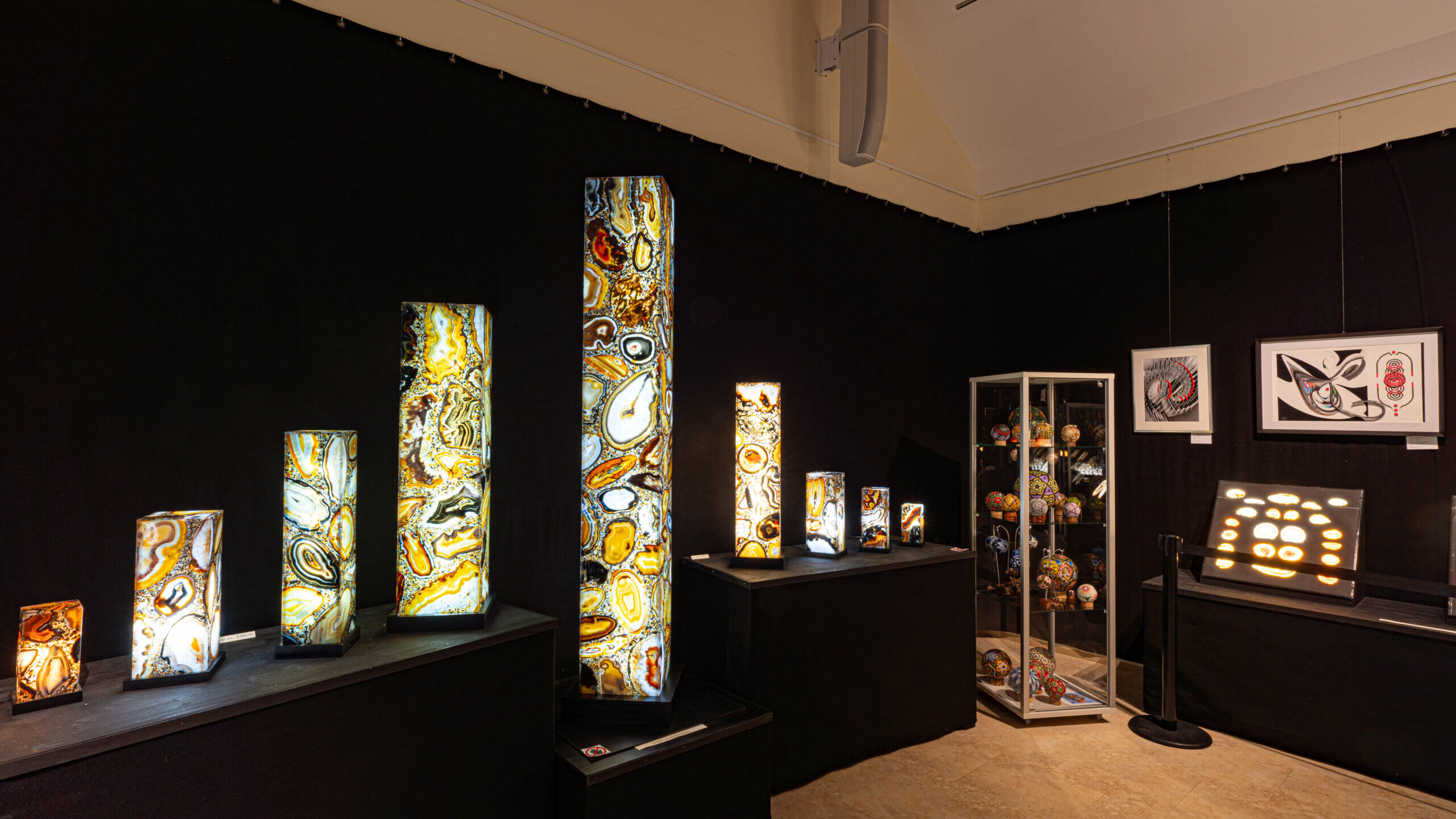 Ausstellung beleuchteter Steinsäulen in Galerie mit abstrakter Kunst und dekorativen Objekten im Hintergrund.