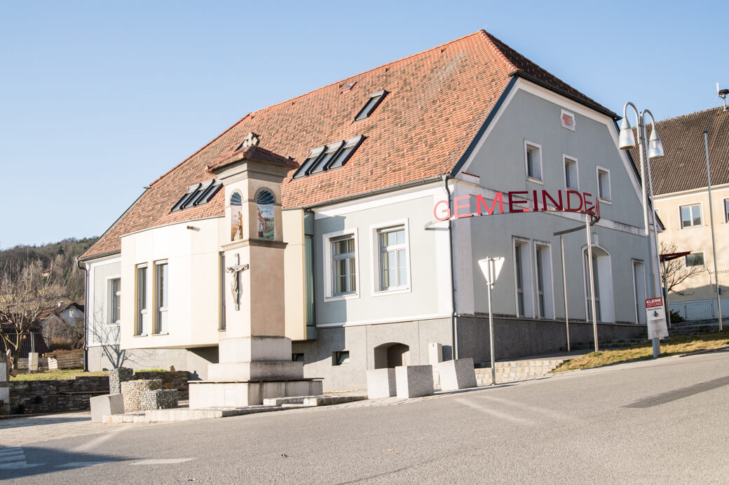 Historisches Gemeindegebäude mit rotem Dach und Jesuskreuz im sonnigen Dorf, seitlich eine Laterne.
