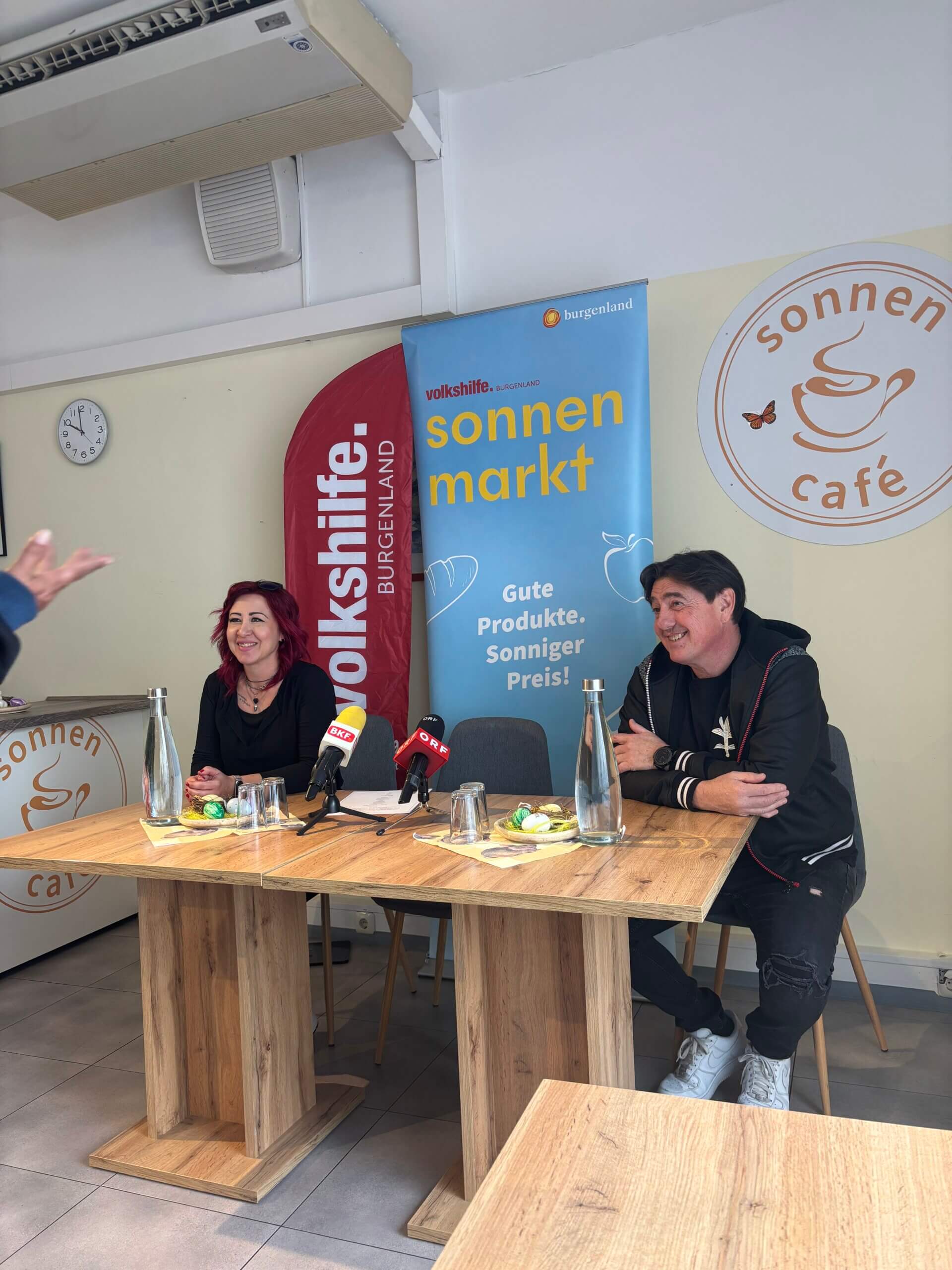 Pressekonferenz im Sonnenmarkt: Zwei Personen am Tisch mit Mikrofonen. Banner von Volkshilfe Burgenland im Hintergrund.