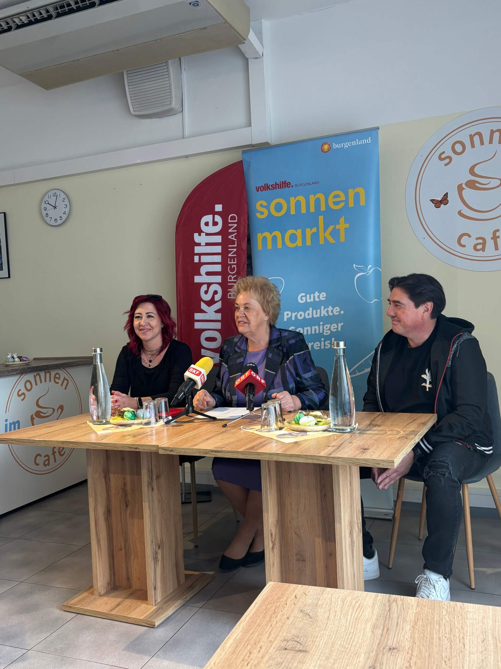 Pressekonferenz im Sonnenmarkt Burgenland, drei Personen sprechen am Tisch, Logos von Volkshilfe und Sonnenmarkt im Hintergrund.