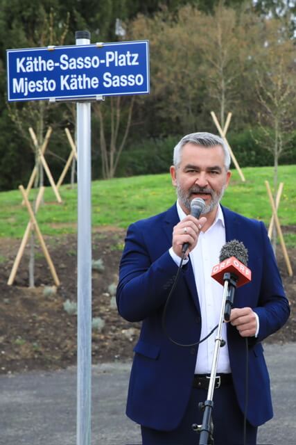 Mann spricht bei Einweihung des Käthe-Sasso-Platzes, Schild im Hintergrund, Außenaufnahme, Mikrofon in der Hand.
