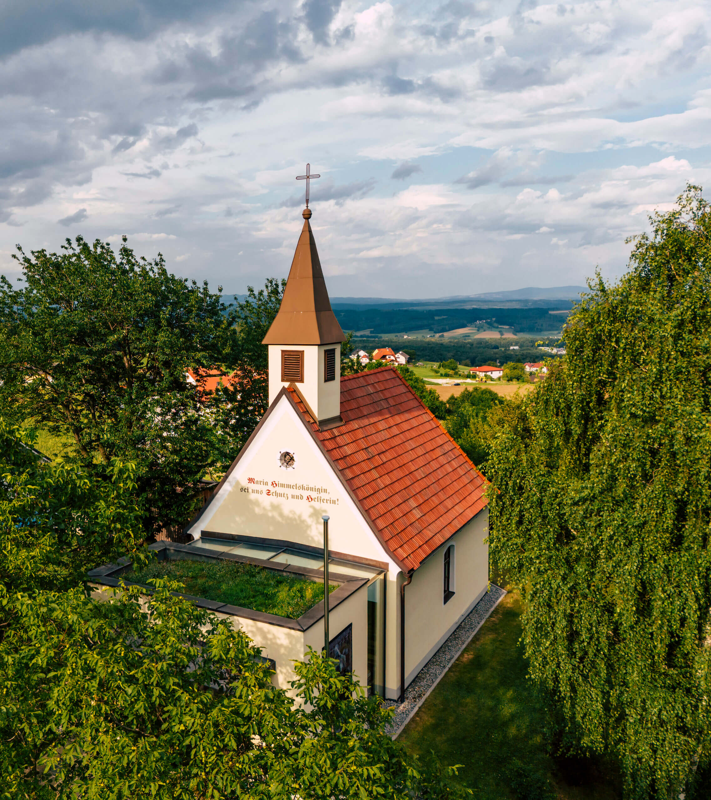 Kleine Kapelle mit rotem Dach in idyllischer Landschaft unter bewölktem Himmel, umgeben von Bäumen.