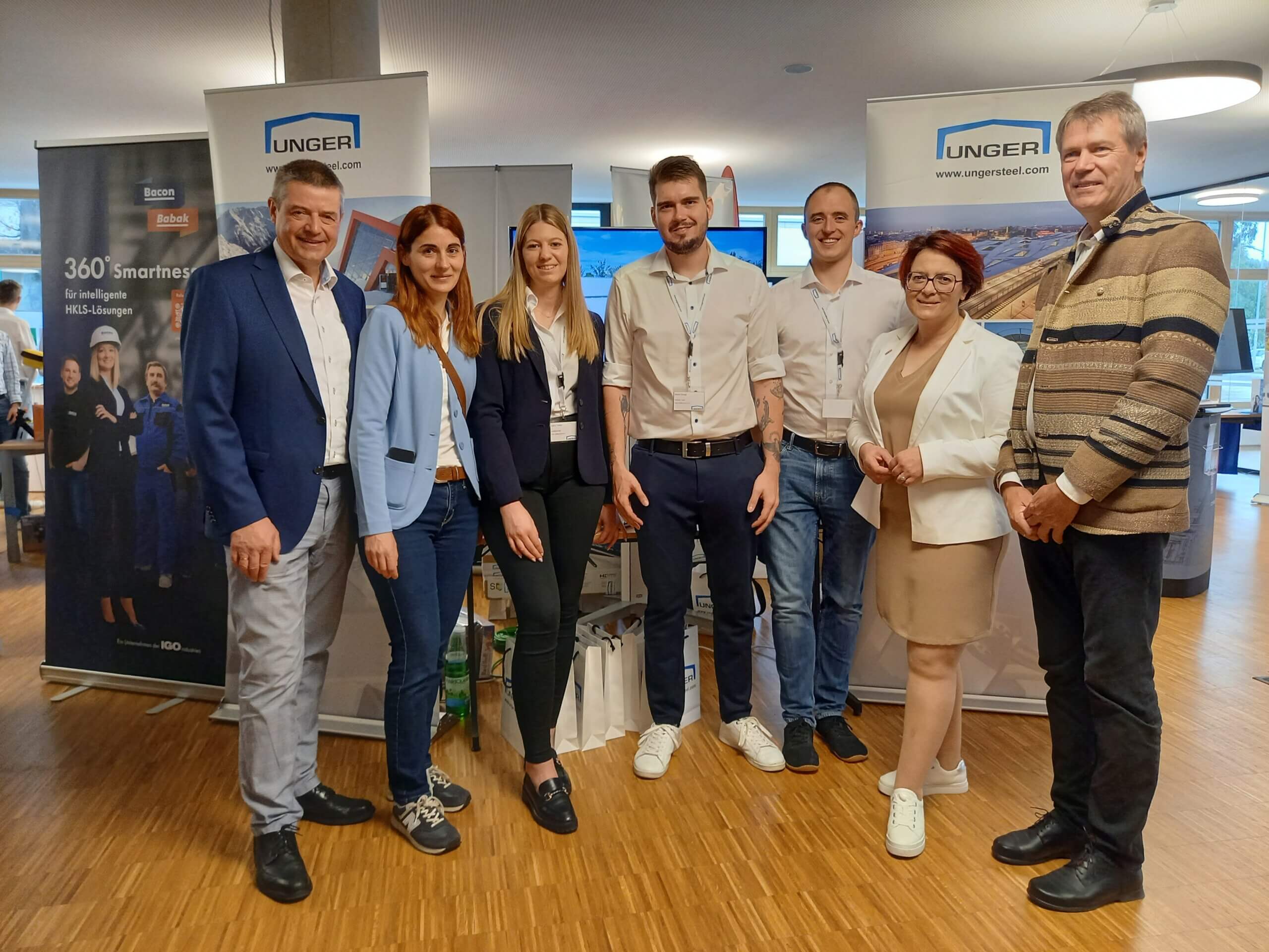 Gruppe von sieben Menschen auf einer Firmenveranstaltung vor zwei Bannern in einem modernen Tagungsraum.