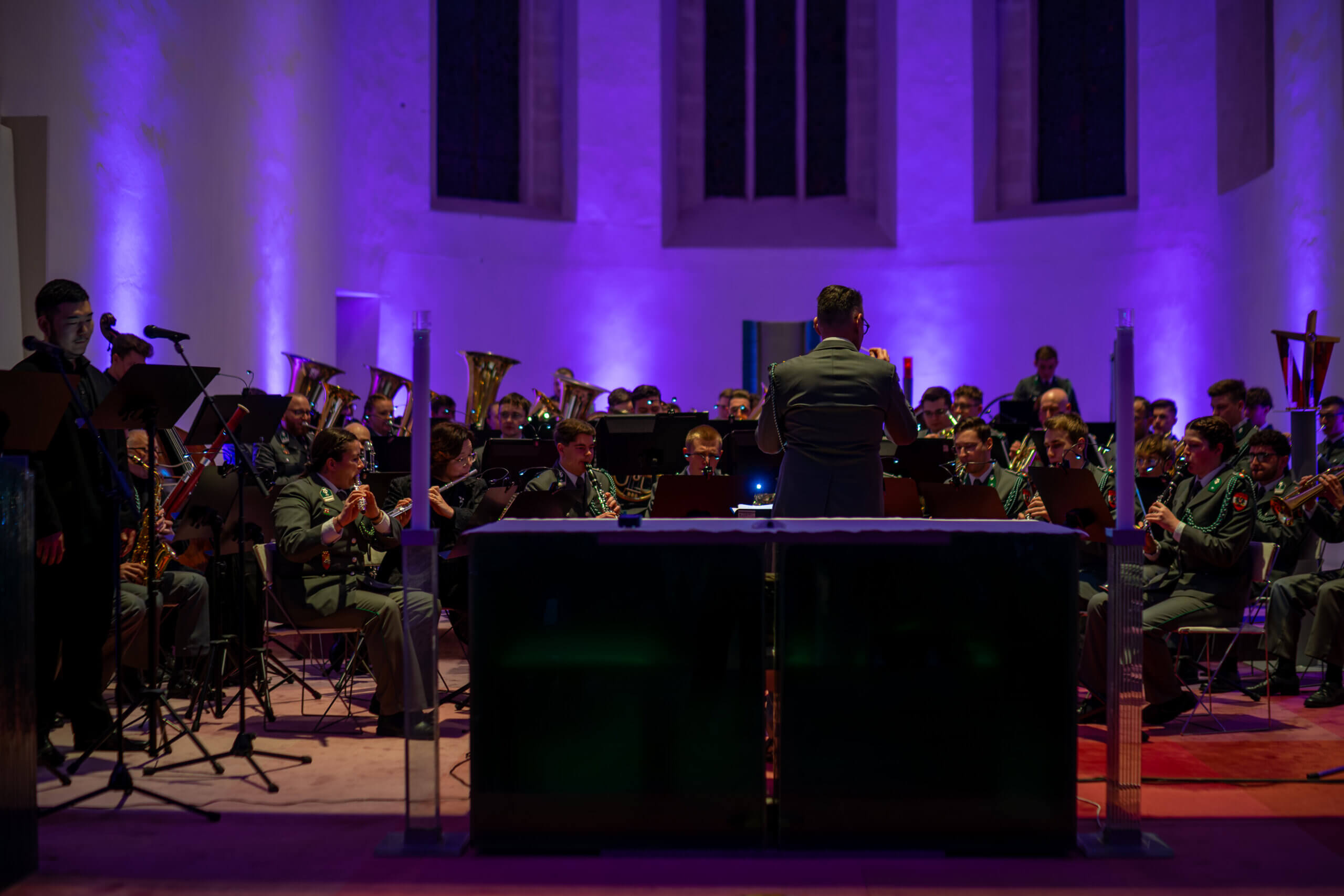 Orchester spielt in beleuchteter Kirche bei Konzert, dirigiert von Maestro; Musiker in Uniformen an Blasinstrumenten.