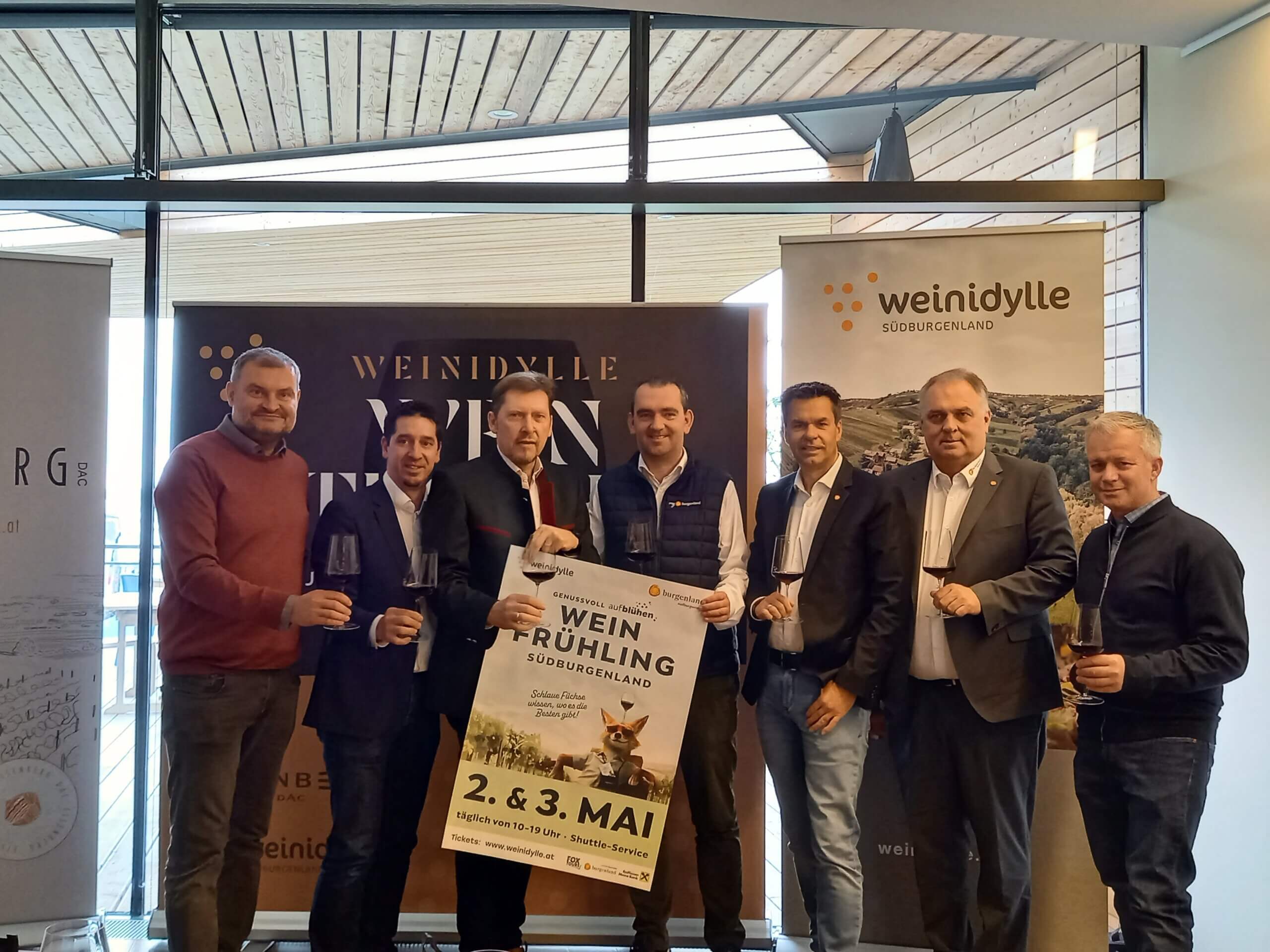 Gruppe von Männern präsentiert Weinfrühling-Event im Südburgenland, hält Weingläser und Plakat hoch.