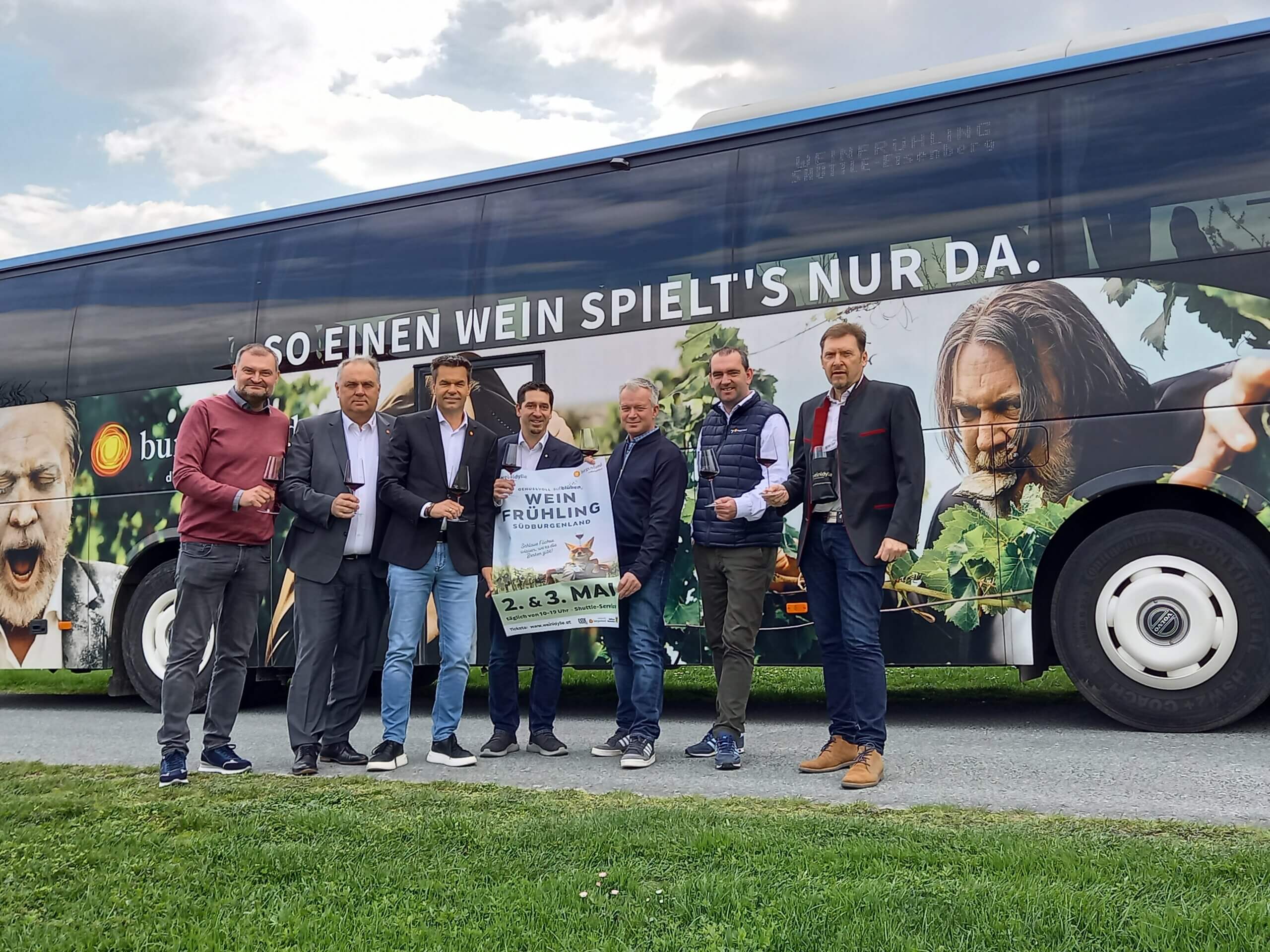 Gruppe von Männern vor einem Bus mit Wein-Werbung, hält ein Plakat für das Weinfestival Südburgenland.