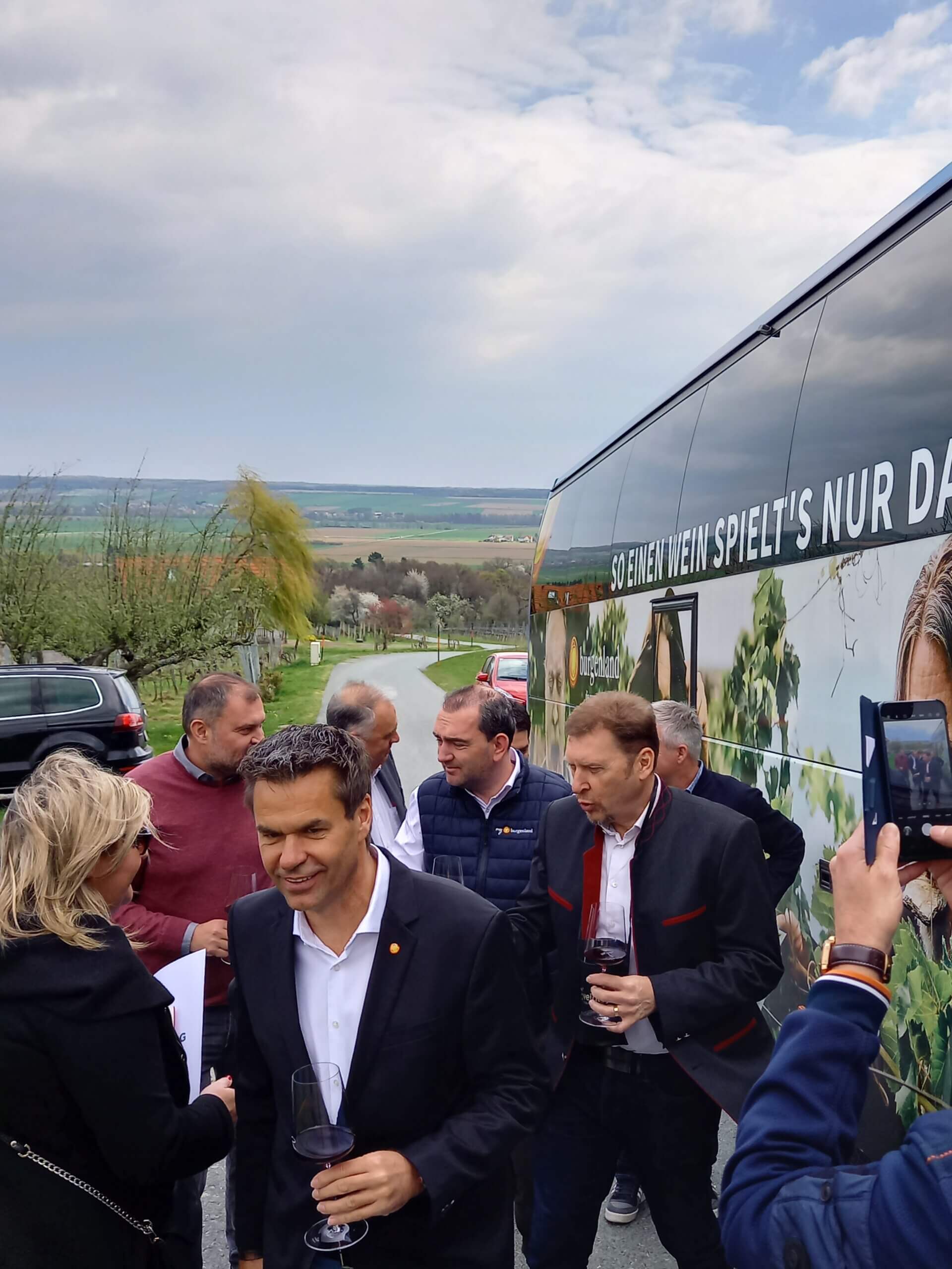 Personengruppe beim Weintasting vor dekoriertem Bus in idyllischer Landschaft.