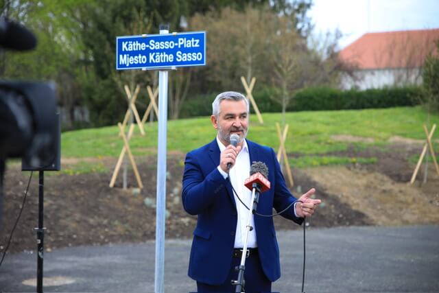 Mann hält Rede neben Schild Käthe-Sasso-Platz im Freien, frühlingshafte Szenerie im Hintergrund.