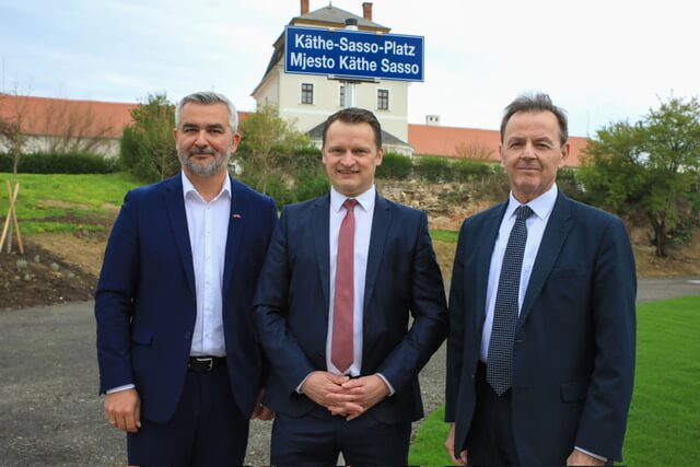 Drei Herren in Anzügen am Käthe-Sasso-Platz, Wien, mit Gebäuden im Hintergrund.