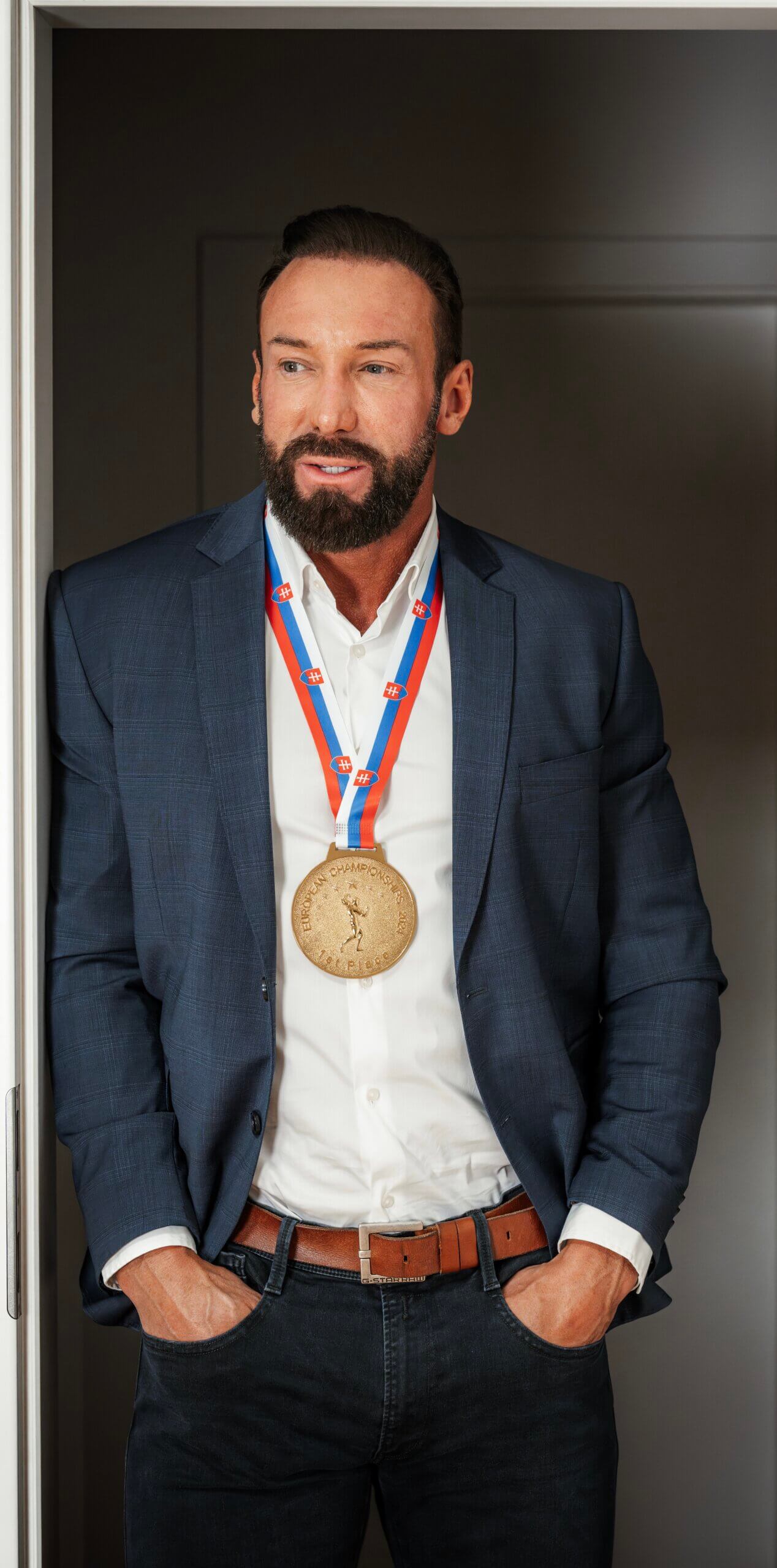 Mann in Anzug mit Medaille, lässig posierend vor einer Tür. Business-Erfolg und Auszeichnung im Fokus.