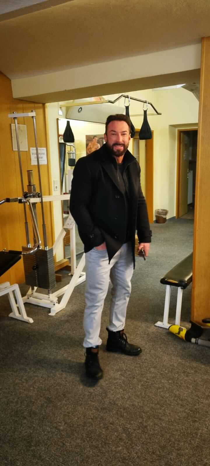 Mann im Fitnessstudio, lächelnd und in schwarzer Jacke, neben Trainingsgeräten, Innenraumansicht.