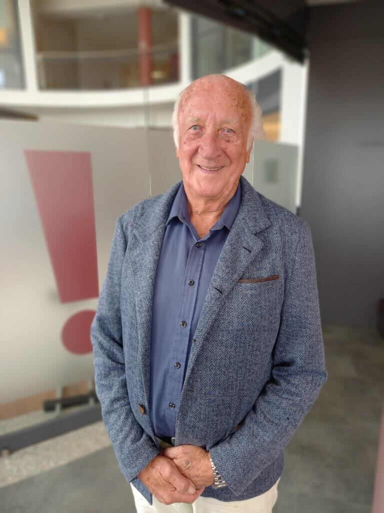 Alfred Nöhrer©Nicole Matsch Älterer Mann in blauem Blazer lächelt in moderner Umgebung, Ausrufezeichen im Hintergrund.