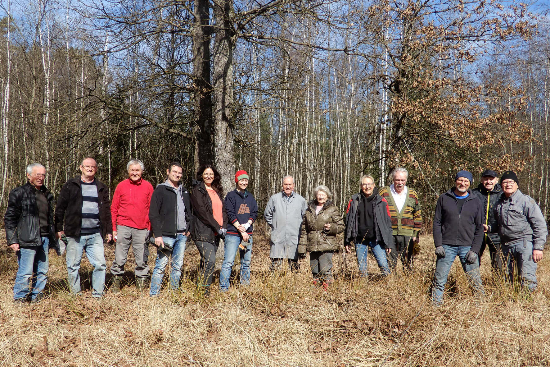 Gruppe von Menschen in Freizeitkleidung posiert lächelnd im Freien vor einem Waldgebiet an einem sonnigen Tag.