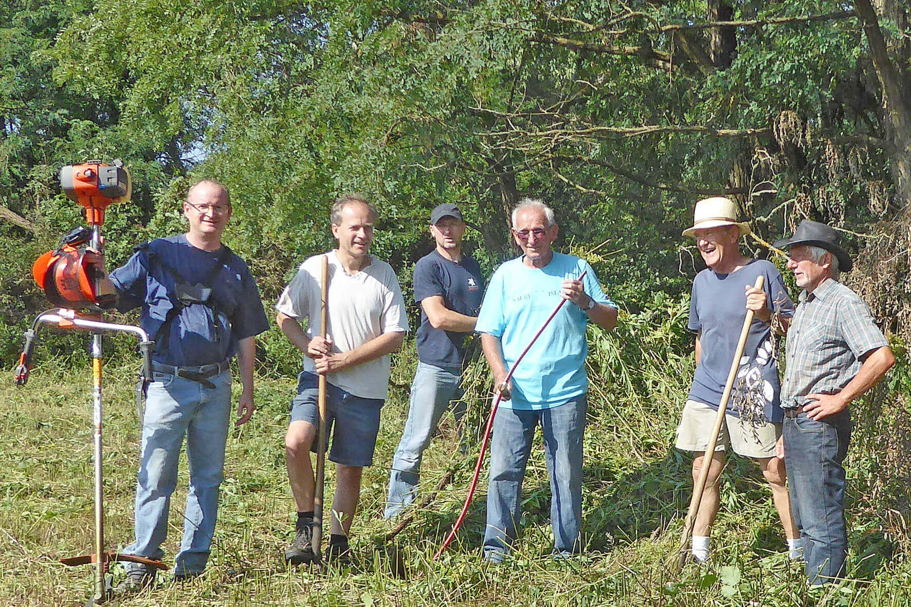 Gruppe von Männern bei Gartenarbeit mit Werkzeugen bei sonnigem Wetter, umgeben von Bäumen und Grünflächen.