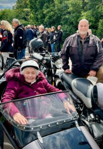 Ältere Frau im Beiwagen eines Motorrads, lachend, umgeben von Motorradfahrern bei einer Veranstaltung im Freien.