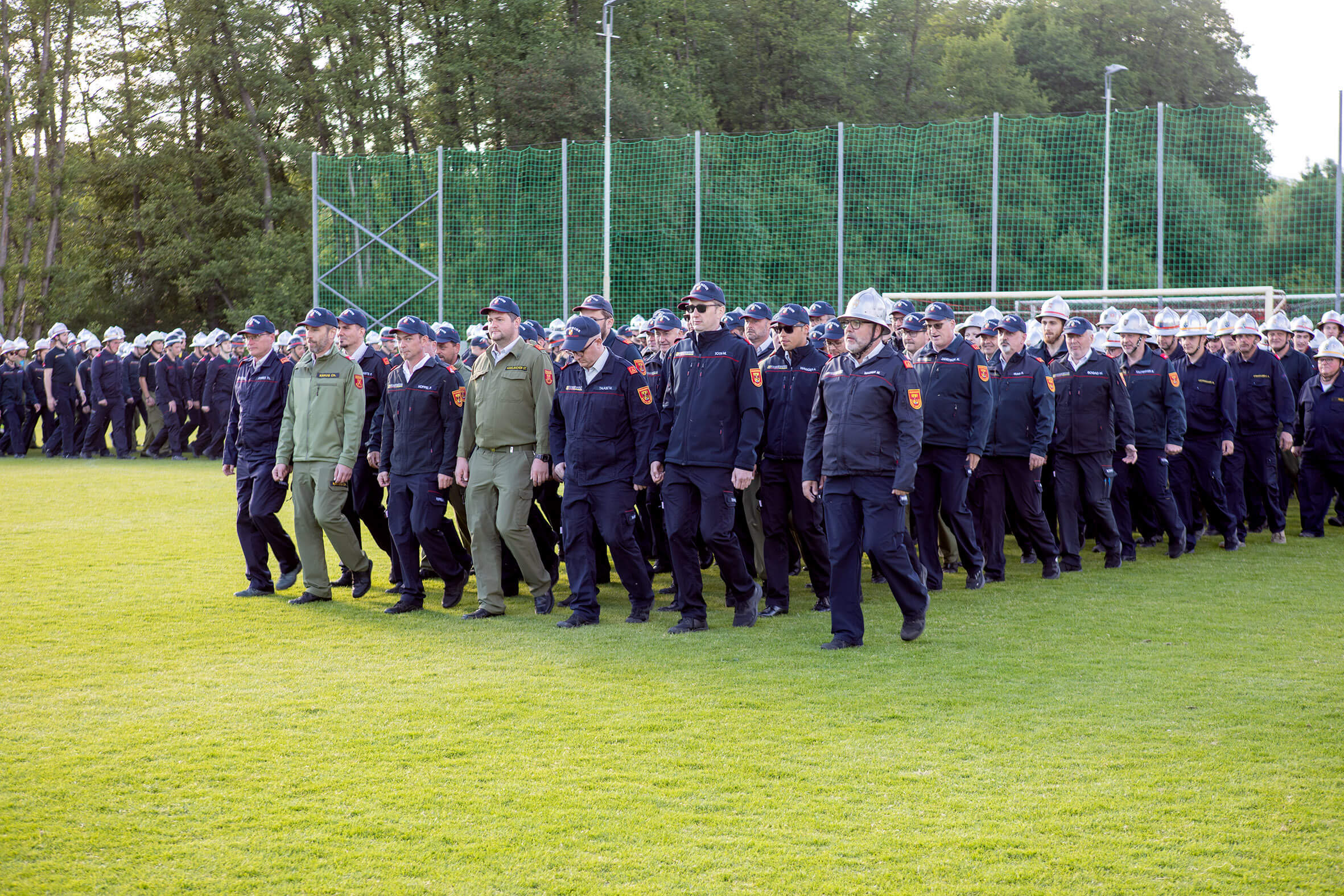 Gruppe von Feuerwehrleuten marschiert in Uniform auf einem Sportplatz, angetreten zu einer feierlichen Veranstaltung.