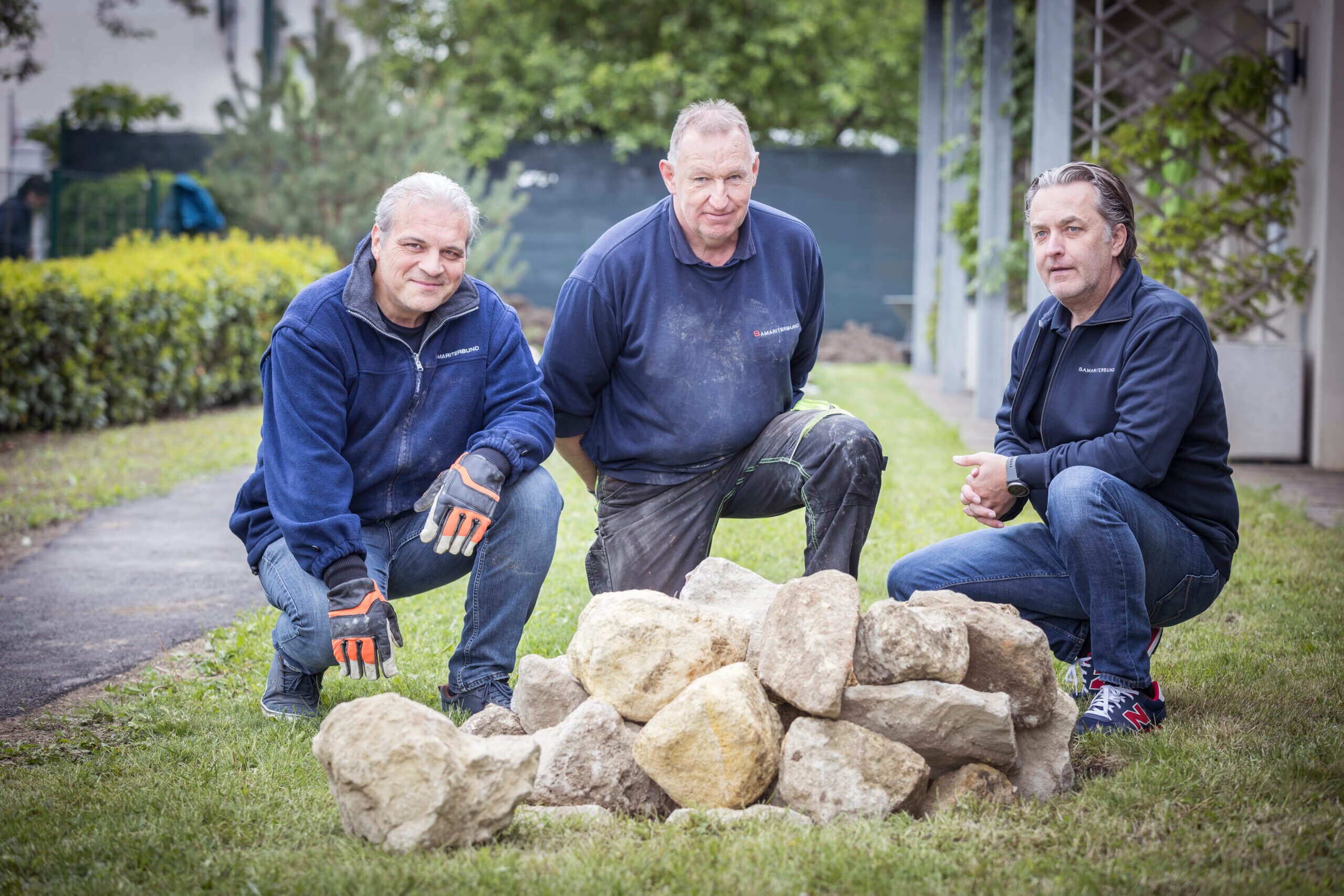 Drei Männer in Arbeitskleidung knien auf Rasen, inspizieren Steinaufbau, im Hintergrund Bäume und Gebäude zu sehen.