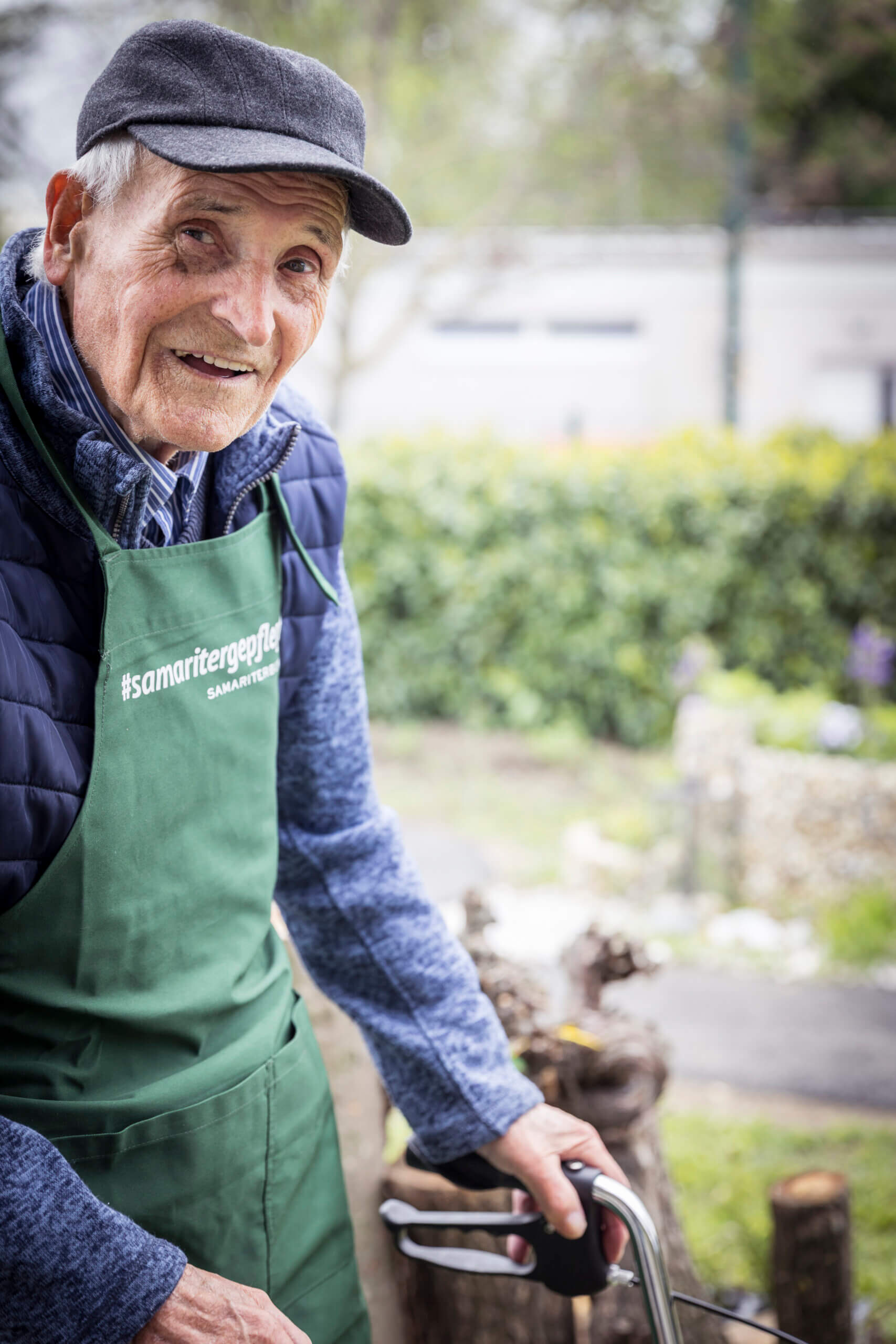 Älterer Mann im Grünen, lächelnd mit Schürze, steht draußen mit Rollator; Thema: Senioren, Gesundheit, Gartenarbeit.