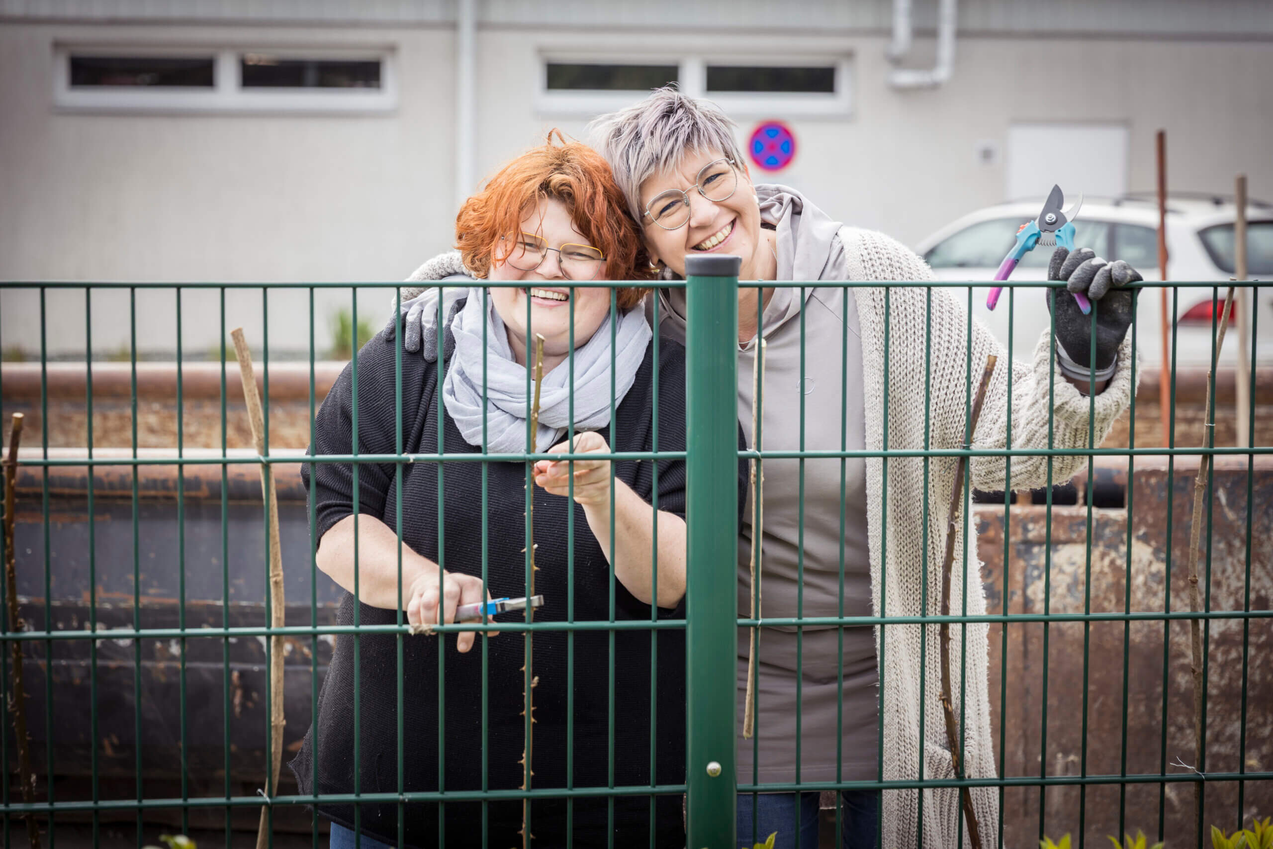 Zwei lächelnde Frauen schneiden hinter einem Zaun Pflanzen mit einer Gartenschere.