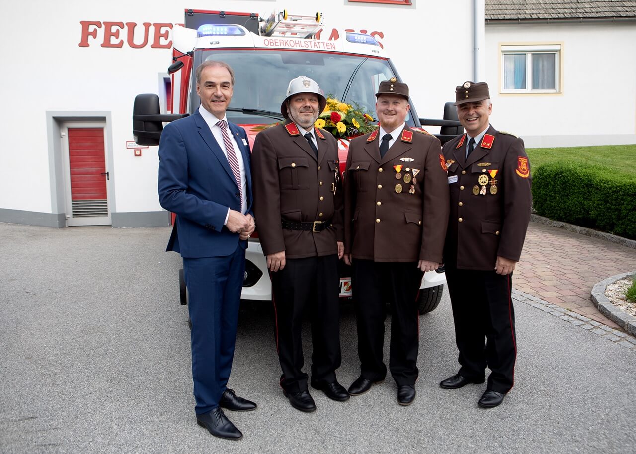 Feuerwehrleute vor einem Einsatzfahrzeug in Oberkohlstätten bei einer offiziellen Veranstaltung, lächelnd in Uniform.