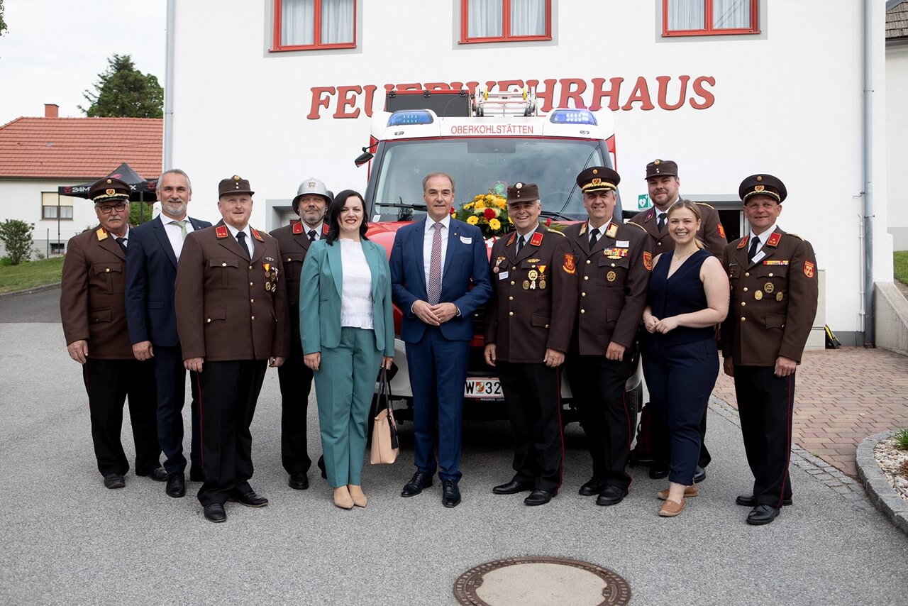 Gruppe vor Feuerwehrhaus Oberkohlstätten, versammelte Feuerwehrleute und Gäste lächeln vor einem Einsatzfahrzeug.