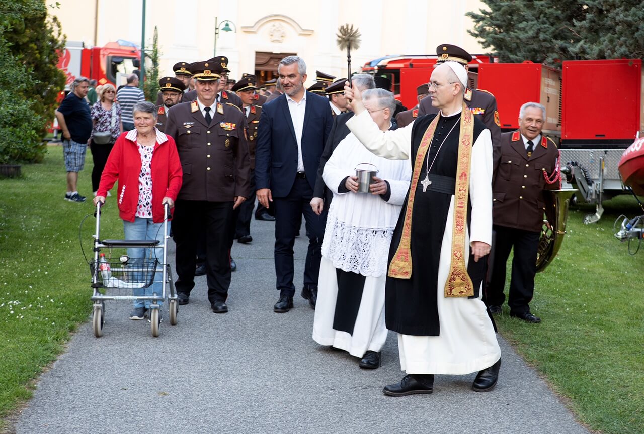 Kirchliche Segnung mit Feuerwehr und Gemeindemitgliedern auf grünem Gelände bei Prozession.