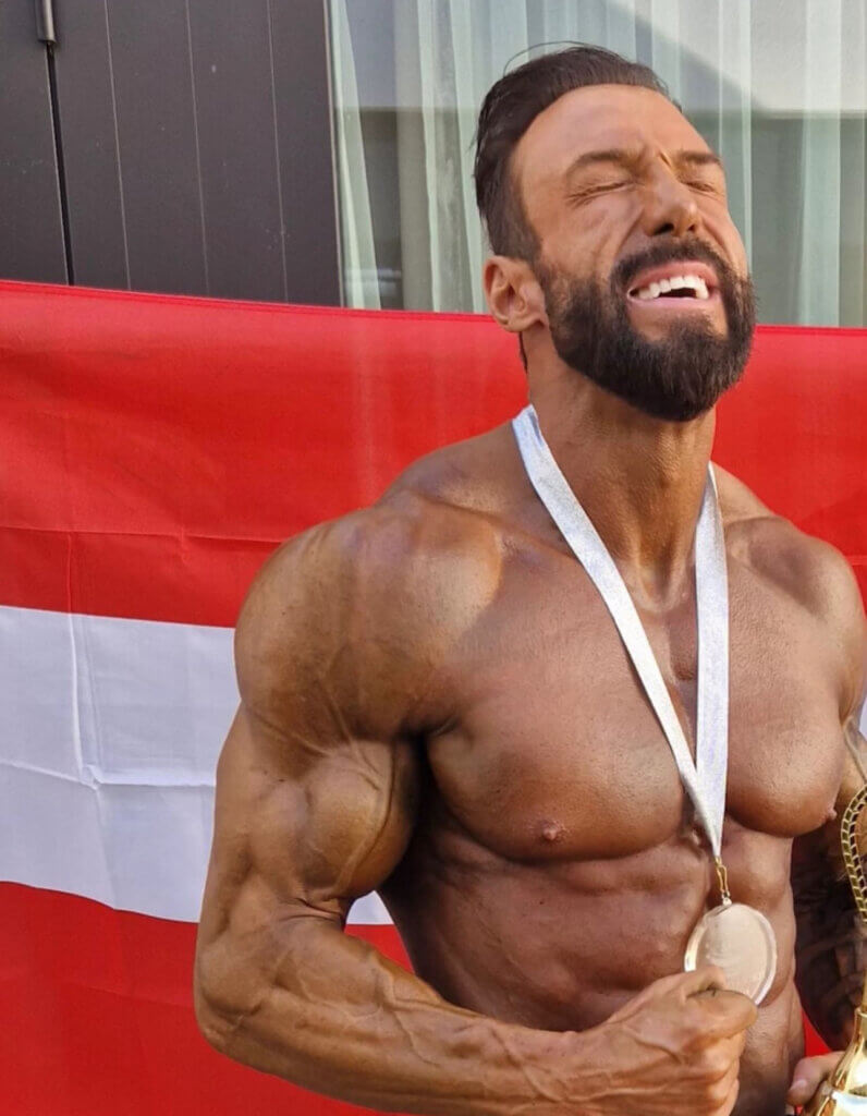 Fuchs 6©Markus Fuchs_b Kraftvoller Bodybuilder mit Medaille vor rotem Hintergrund, zeigt Muskulatur und Stolz auf seine sportliche Leistung.