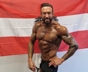 Bodybuilder posiert stolz vor der österreichischen Flagge bei einem Wettbewerb. Topform und Muskeldefinition.
