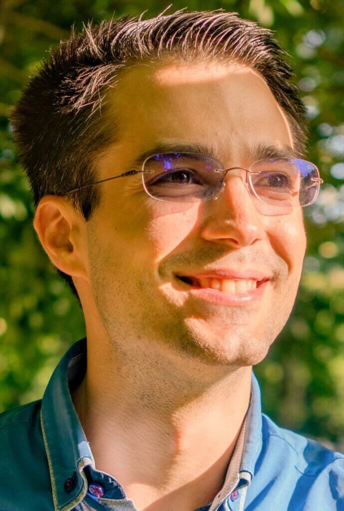 Haider-768x1141 Lächelnder Mann mit Brille im Sonnenlicht, blaues Hemd, grüner Hintergrund. Optimistisch und freundlich.