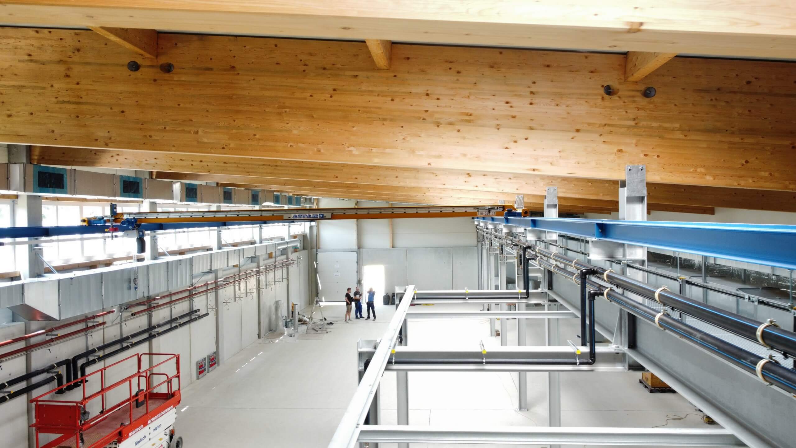 Fabrikhalle mit Holzdecken und Metallstrukturen, Ingenieure besprechen Projekt unter moderner Hebeanlage.