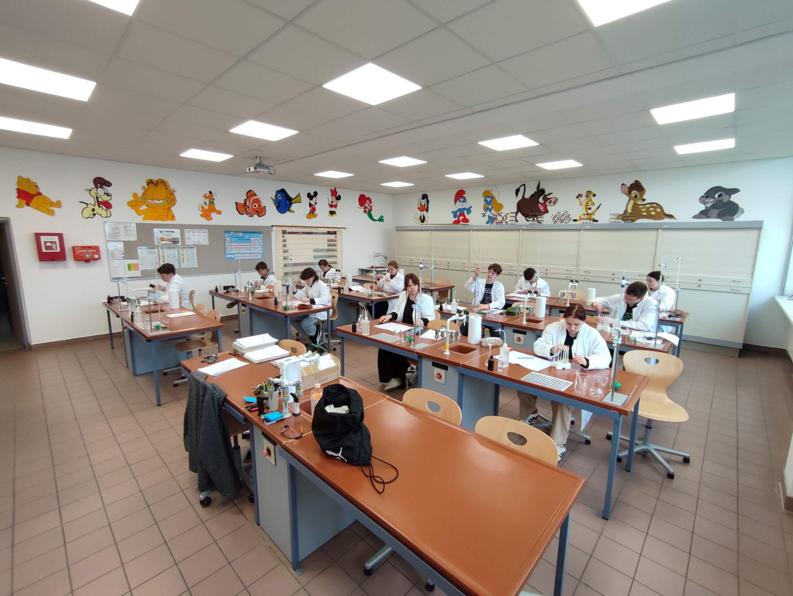 Schüler in weißen Kitteln arbeiten konzentriert in einem farbenfrohen Chemie-Klassenzimmer mit Wanddekorationen.