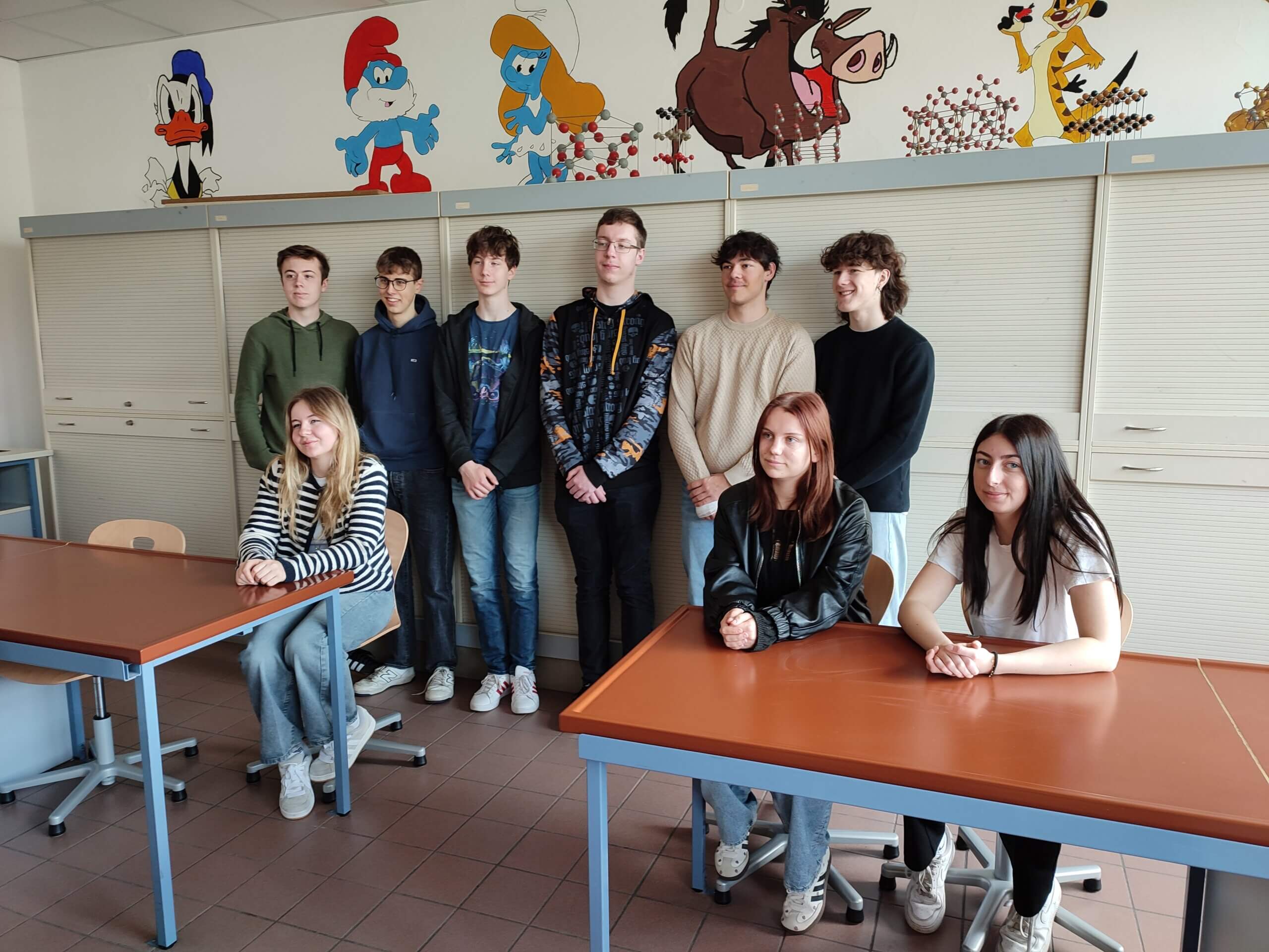 Eine Gruppe von Schülern posiert in einem Klassenzimmer mit bunten Wandbildern im Hintergrund.