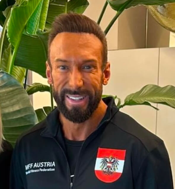 Ein Mann in Sportjacke mit WFF Austria-Logo lächelt vor Pflanzen.