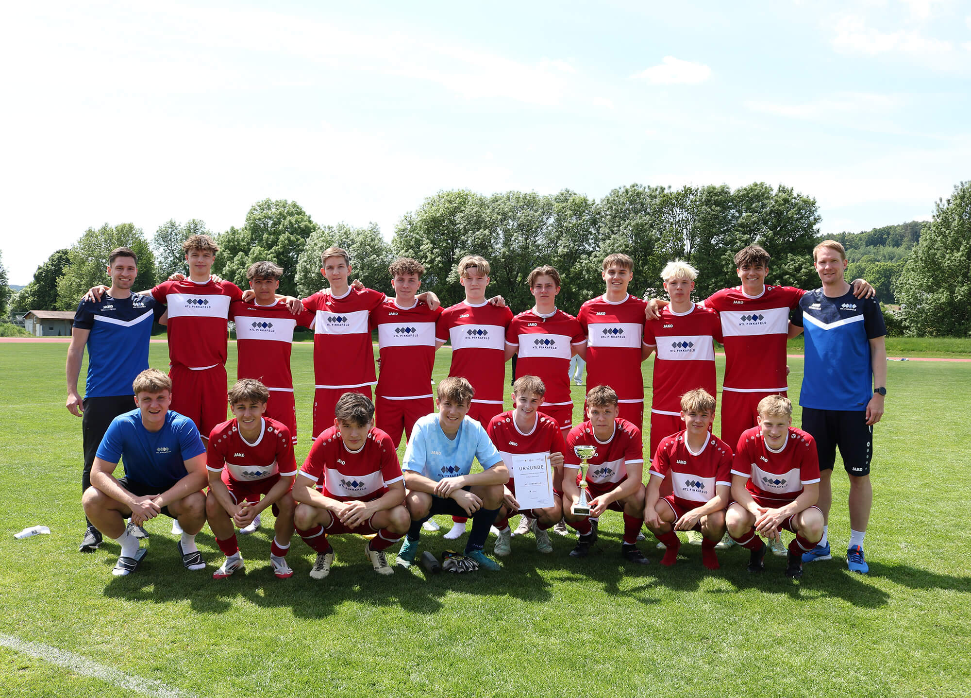 Jugend-Fußballteam in roter Sportkleidung posiert mit Pokal und Urkunde auf grünem Rasen, bei sonnigem Wetter.