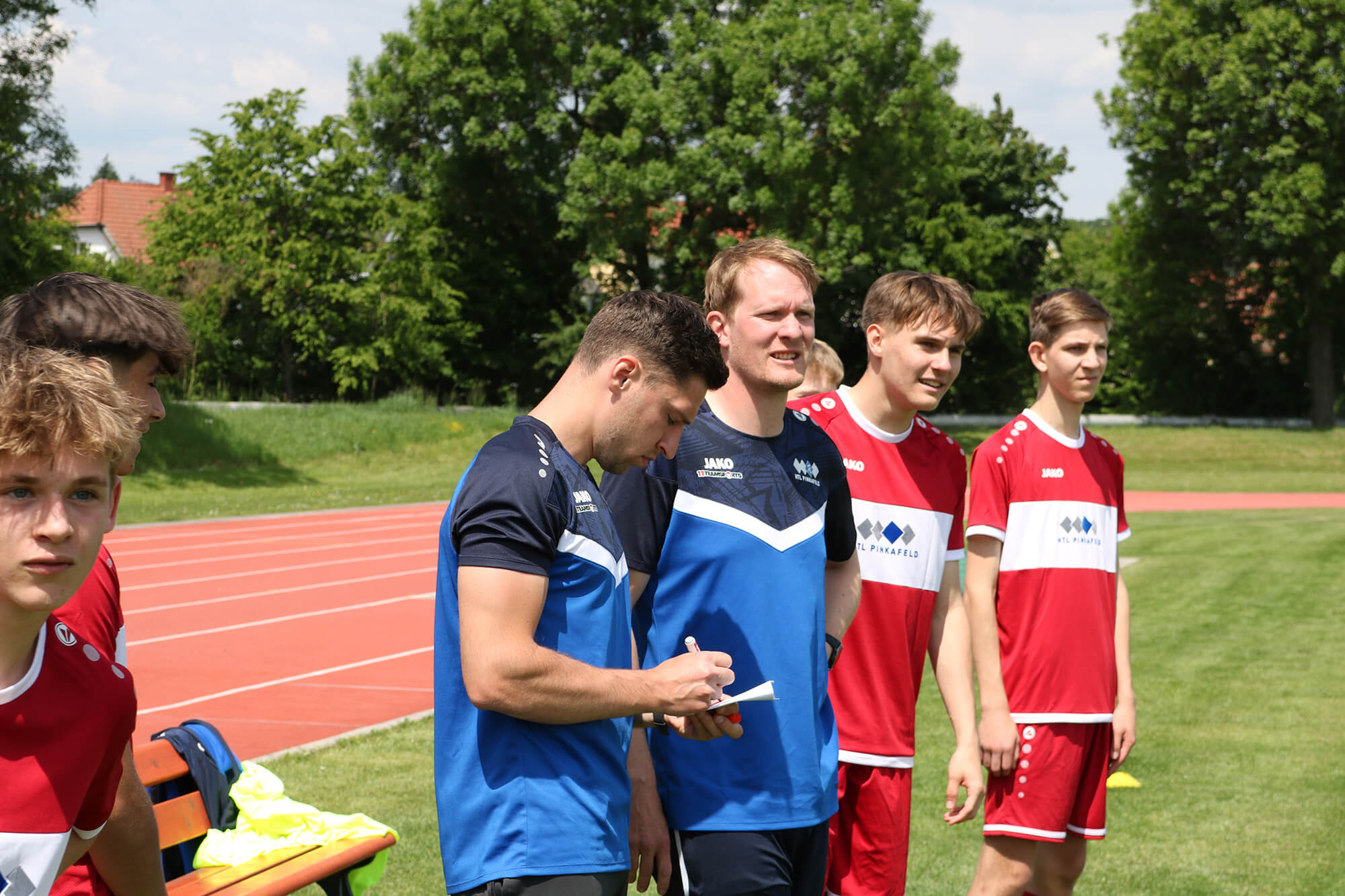 Jugend-Fußballtraining im Freien mit Spielern und Trainer, der Notizen macht, Sommerwetter, grüne Sportanlage.